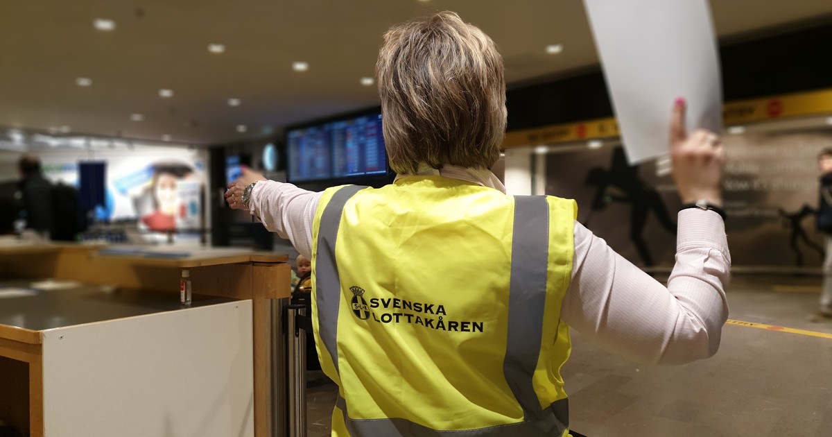 Kvinna bär en reflexväst med texten Svenska Lottakåren. Hon håller ett papper i höger hand och pekar med vänster hand.