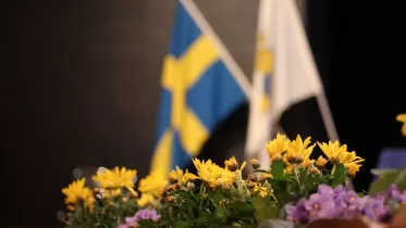 Fotografi av en kruka med gula blommor med två flaggor i bakgrunden, dels den blågula svenska flaggan och sedan standaret för Svenska Lottakåren