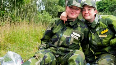 två tjejer sitter på marken och håller om varandra. De tittar leedne in i kameran.