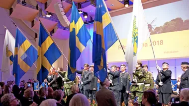 Bild från Riksstämman 2018 som genomfördes genom möte på fysisk plats, lottor som håller svenska flaggan och Lottakårens flagga i förgrunden och mötets podium i bakgrund. I fronten syns lite av publiken som ser upp mot scenen.