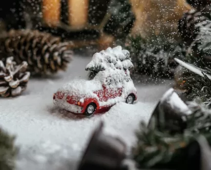 Bild på ett miniatyrlandskap, inzoomat på en översnöad röd leksaksbil med en julgran liggande på taket